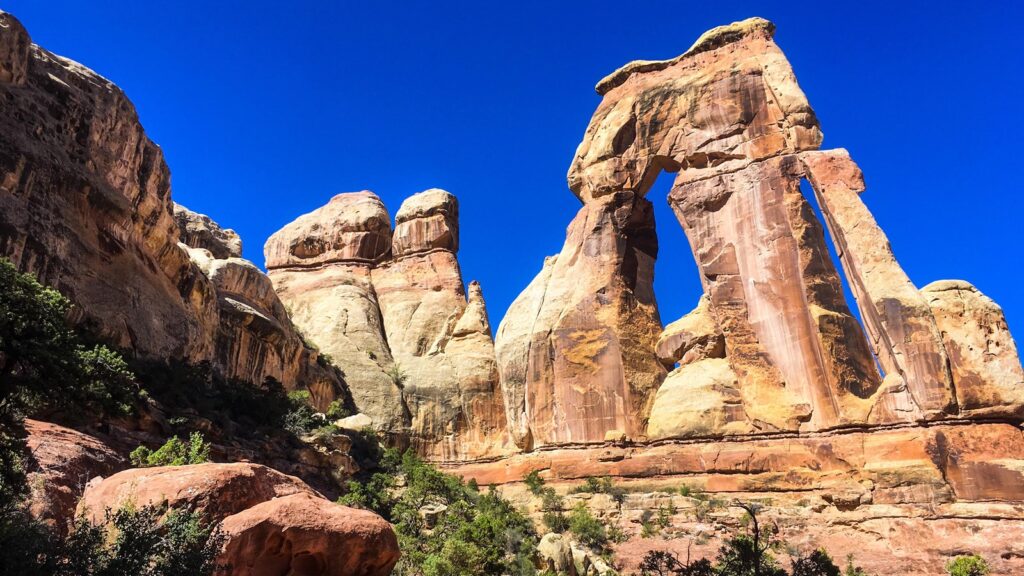 Druid Arch in Canyonlands, a massive sandstone formation with two tall vertical pillars supporting a broad horizontal capstone, viewed from below against a deep blue sky with red sandstone towers nearby.