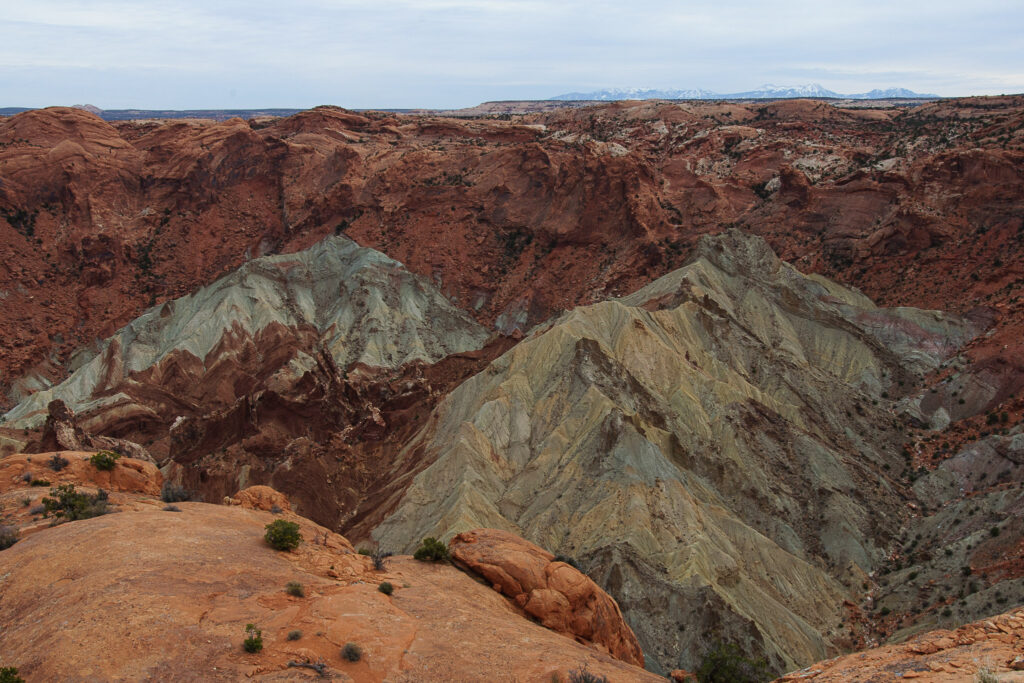Overlook of Upheaval Dome in Canyonlands, showing two eroded blue-gray and tan mounds rising from a canyon, ringed by red sandstone, with snow-capped mountains on the distant horizon.
