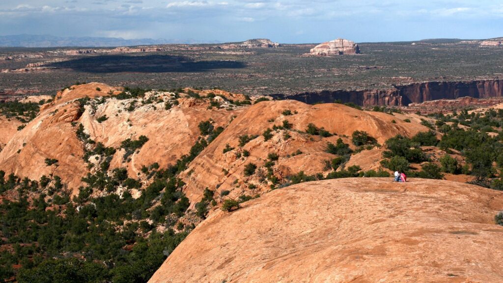 Two hikers rest atop Whale Rock's broad salmon-colored slickrock surface in Canyonlands, with sweeping views of desert plateau, canyon rims, and a white mesa formation beyond.
