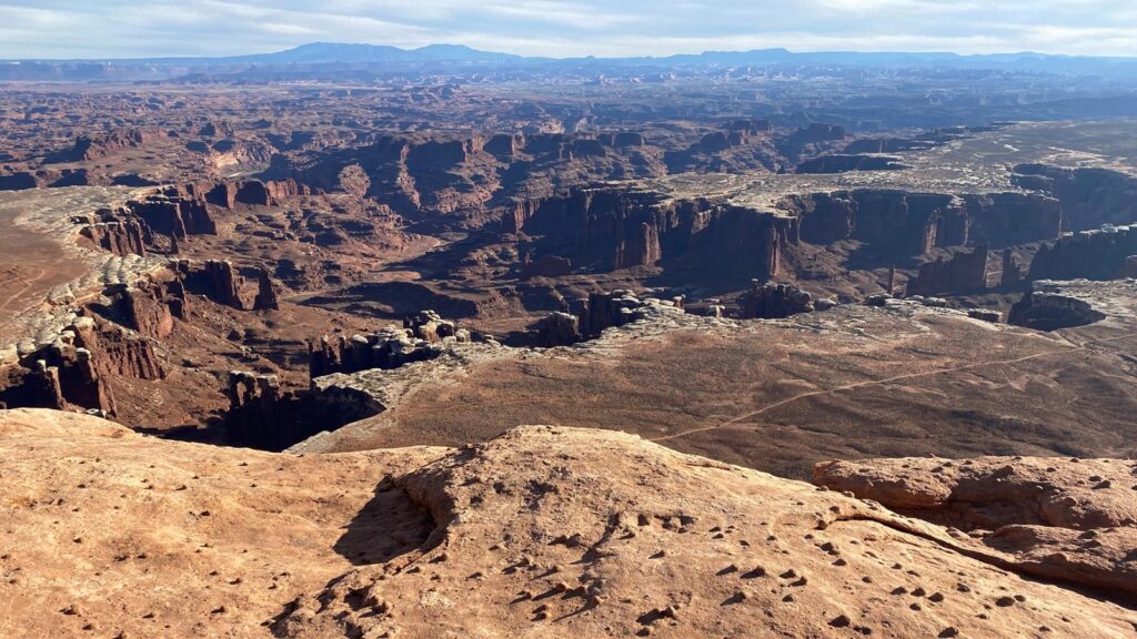 View from White Rim Overlook at Canyonlands, where a smooth sandstone ledge drops sharply into a vast expanse of red mesas, buttes, and deep canyon systems, with faint blue mountains on the horizon.