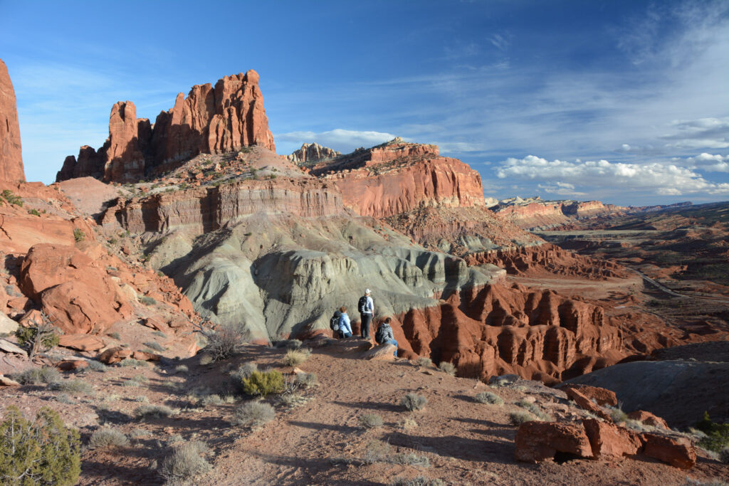 Three hikers stand on a red-rock overlook at Capitol Reef facing a cluster of tall sandstone spires rising above banded hillsides and a broad desert valley.