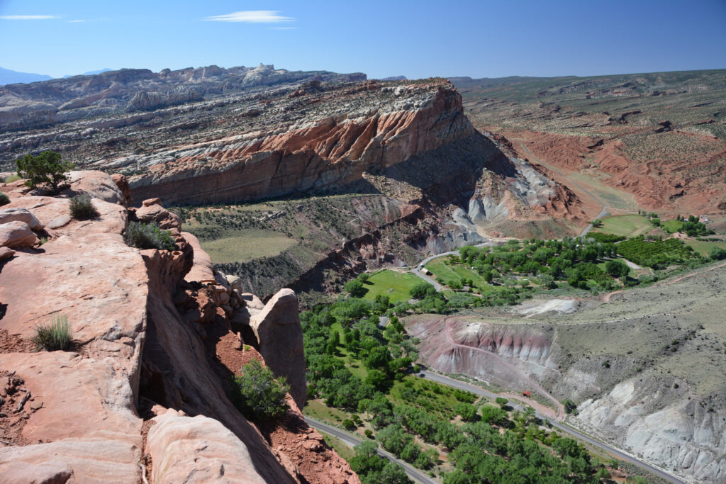View from the Rim Overlook at Capitol Reef: layered red sandstone cliffs drop into a canyon, with a winding road, green valley, and a flat-topped mesa of multicolored strata stretching toward a clear blue horizon.