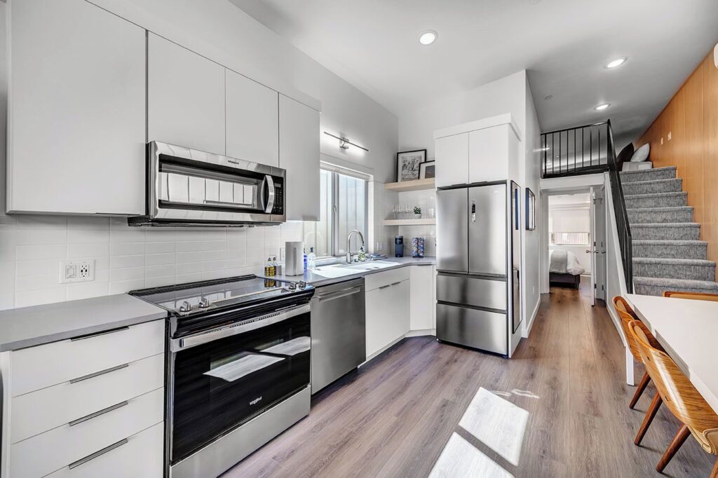Galley kitchen at The Riv in La Verkin, UT, with white flat-front cabinets, stainless steel Whirlpool appliances, subway tile backsplash, and hardwood-style flooring, with a loft staircase visible at right.