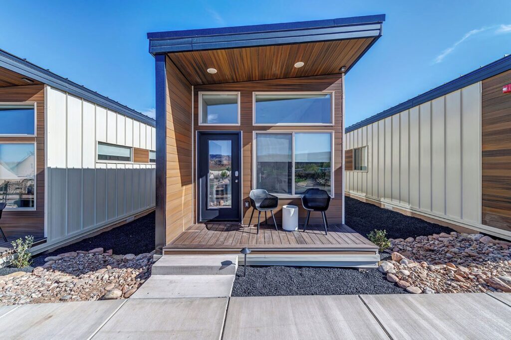 Front exterior of a modern tiny cabin at The Riv in La Verkin, UT, with warm wood siding, a flat canopy, black glass entry door, and a small deck with two chairs set amid desert gravel landscaping.