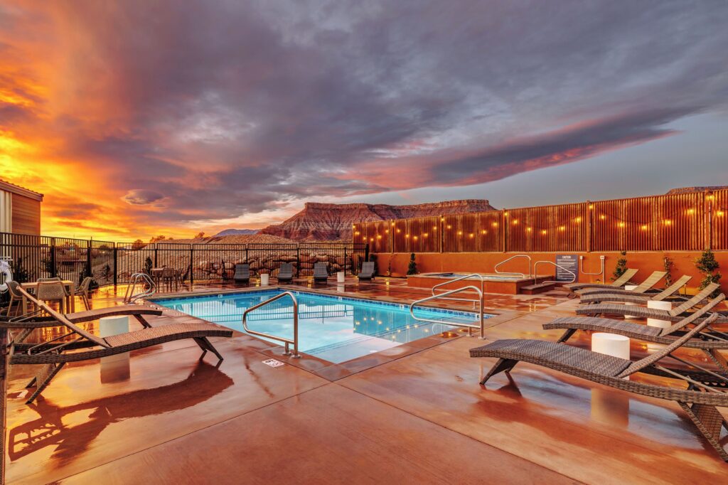 Illuminated outdoor pool and hot tub at The Riv in La Verkin, UT, with string-lit slatted fencing, wicker lounges, and a red rock mesa silhouetted against a vivid orange and purple sunset.