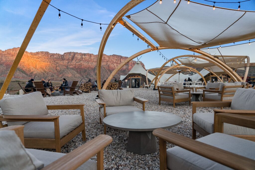 Outdoor lounge at Under Canvas Zion with teak chairs and loveseats under canvas shade sails and Edison lights, guests gathered around a fire pit, with red sandstone cliffs glowing at dusk behind white glamping tents.