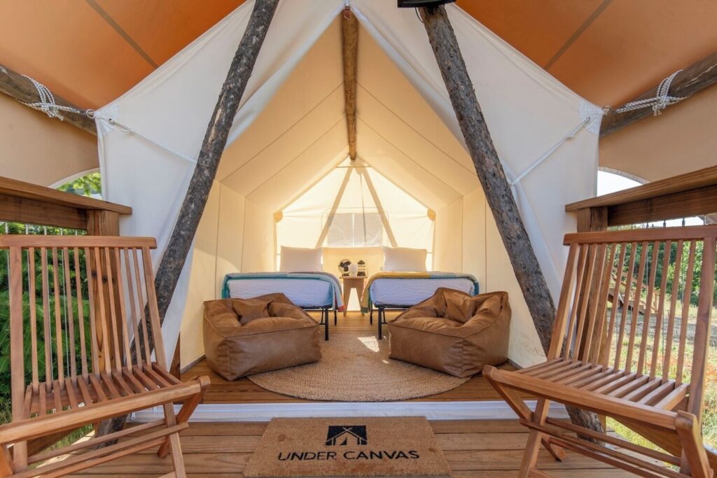 Looking inward through the open canvas flap of an Under Canvas Zion glamping tent, two twin beds with white bedding face brown leather bean bag chairs on a jute rug, with rustic log poles framing the interior.