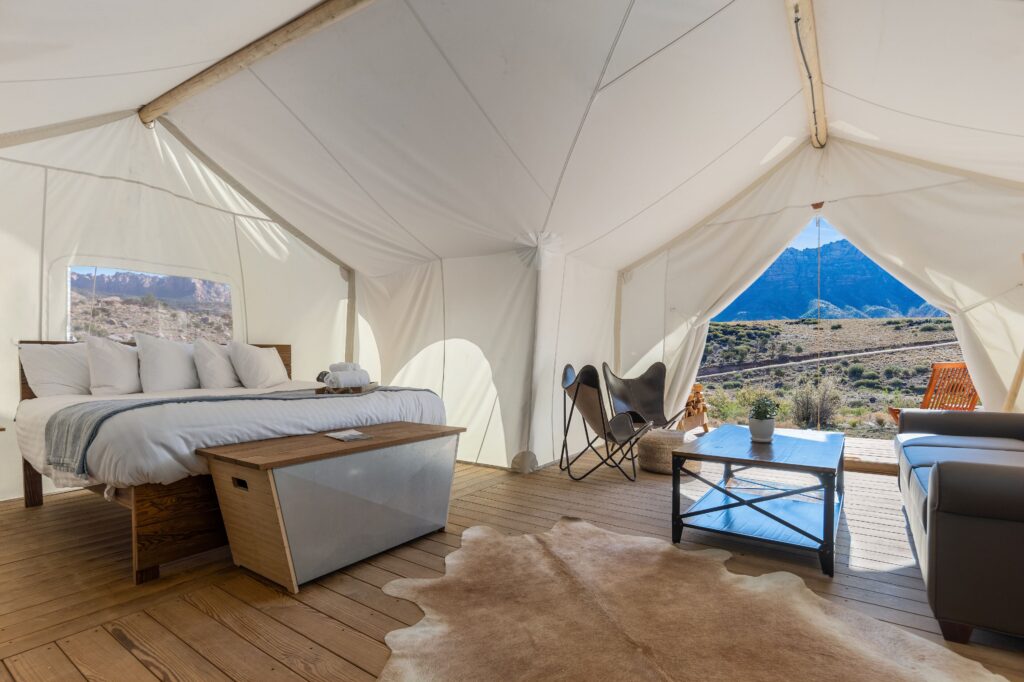 Inside an Under Canvas Zion glamping tent in La Verkin, UT, a king bed with white linens and a cowhide rug face an open entrance framing red rock canyon views beyond.