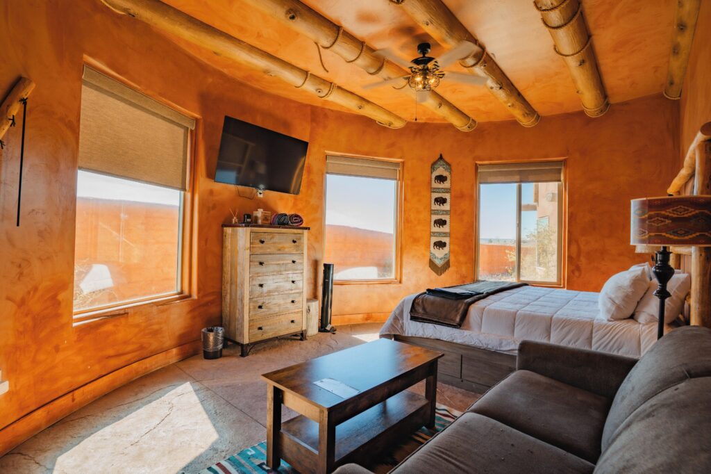 Interior of a glamping suite at Zion White Bison Glamping & RV Resort in La Verkin, featuring adobe plaster walls, log beam ceiling, queen bed with white bedding, and windows overlooking the desert.