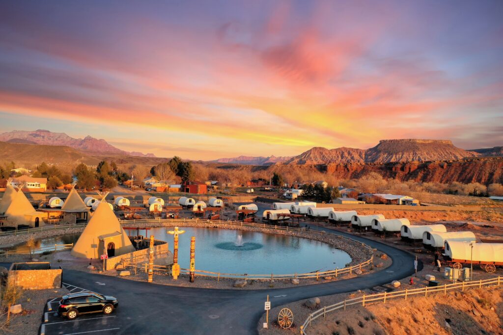 Aerial view of Zion White Bison Glamping & RV Resort near La Verkin, UT at sunset, showing a circular fountain pond, tepee structures, and covered wagon accommodations against red rock mesas under a vivid pink and orange sky.