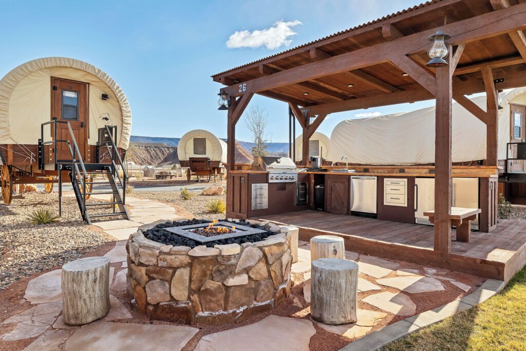 Covered wagon glamping units surround a stone fire pit at Zion White Bison Glamping & RV Resort in La Verkin, Utah, with a pergola-shaded outdoor kitchen on a deck and a desert mesa visible beyond.
