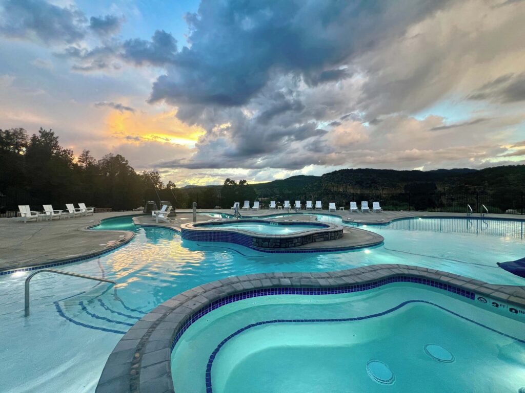 Freeform multi-level pool and spa glowing turquoise at East Zion Resort in Orderville, UT, photographed at sunset beneath dramatic storm clouds with warm orange light over forested hills and rocky ridgelines.