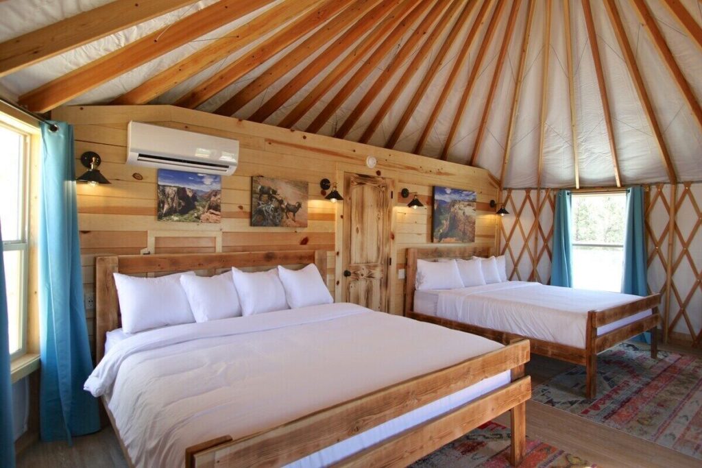 Interior of a glamping yurt at East Zion Resort, with two queen beds, white bedding, teal curtains, and a radiating wood-beam ceiling overhead.