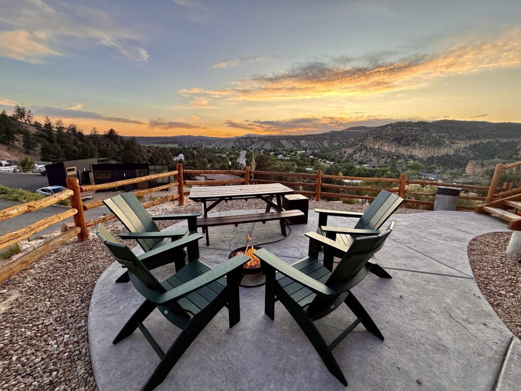 Four dark green Adirondack chairs around a fire pit on a patio at East Zion Resort, with a dramatic golden sunset illuminating canyon and mesa views beyond a log rail fence.