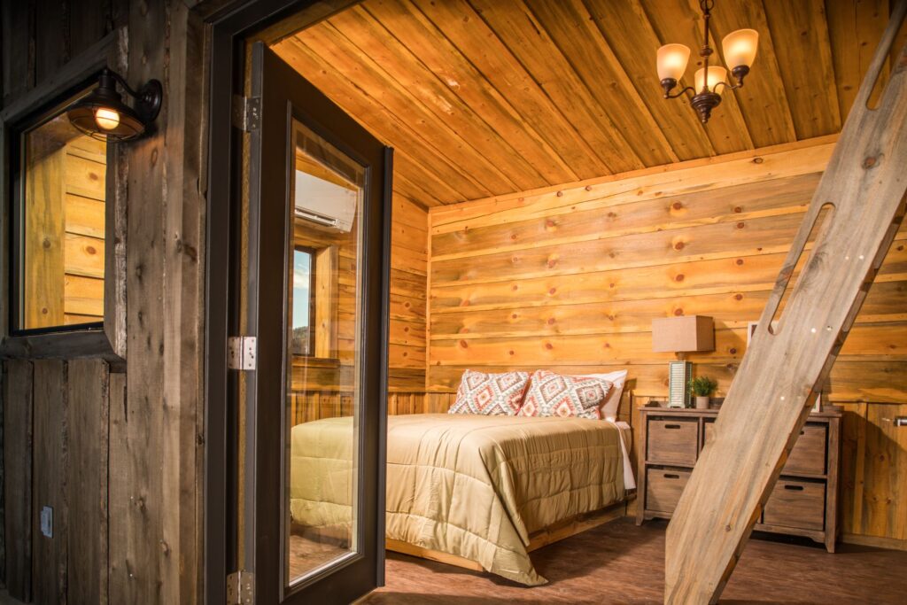 Interior of a Zion Mountain Ranch cabin bedroom with a queen bed, Southwestern-patterned pillows, vaulted wood ceiling, chandelier, and a loft ladder along the wall.