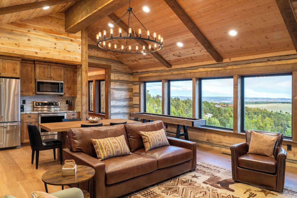 Interior of a Zion Mountain Ranch log cabin great room with vaulted timber-beam ceiling, wagon-wheel chandelier, leather seating, and windows overlooking forested hills and distant mountains.