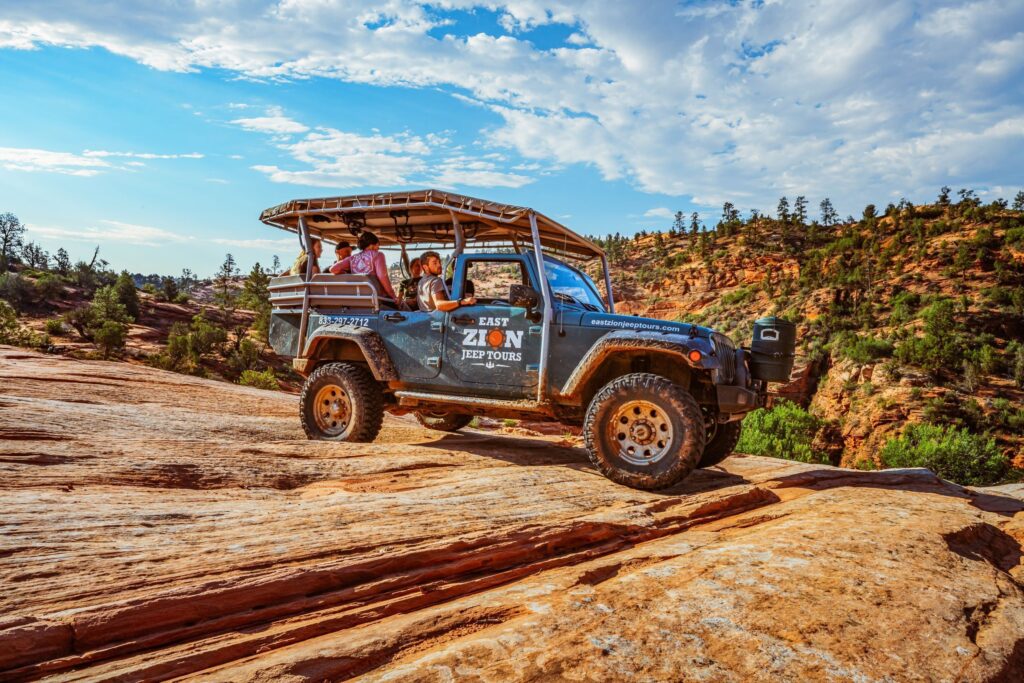 An open-air East Zion Jeep Tours vehicle carries passengers across smooth red slickrock, with sandstone cliffs and scattered pines rising behind it at Zion Ponderosa Resort.