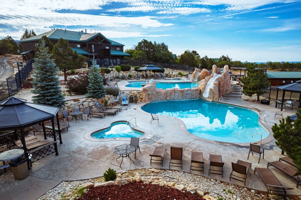 Aerial view of Zion Ponderosa Resort's outdoor pool complex in Orderville, Utah, with a freeform swimming pool, hot tub, rock water slide, and lounge areas beside a log-style lodge building.