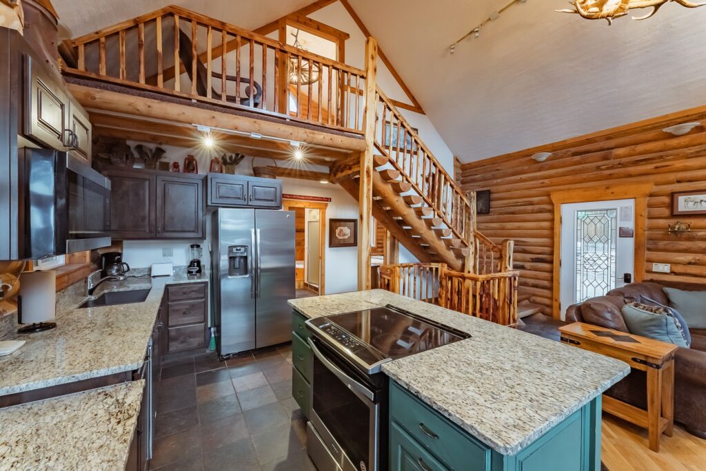 Kitchen and living area inside a Zion Ponderosa Resort rental unit, featuring a teal island, granite countertops, stainless appliances, leather sofa, and a log staircase rising to a lofted upper level under a vaulted ceiling.
