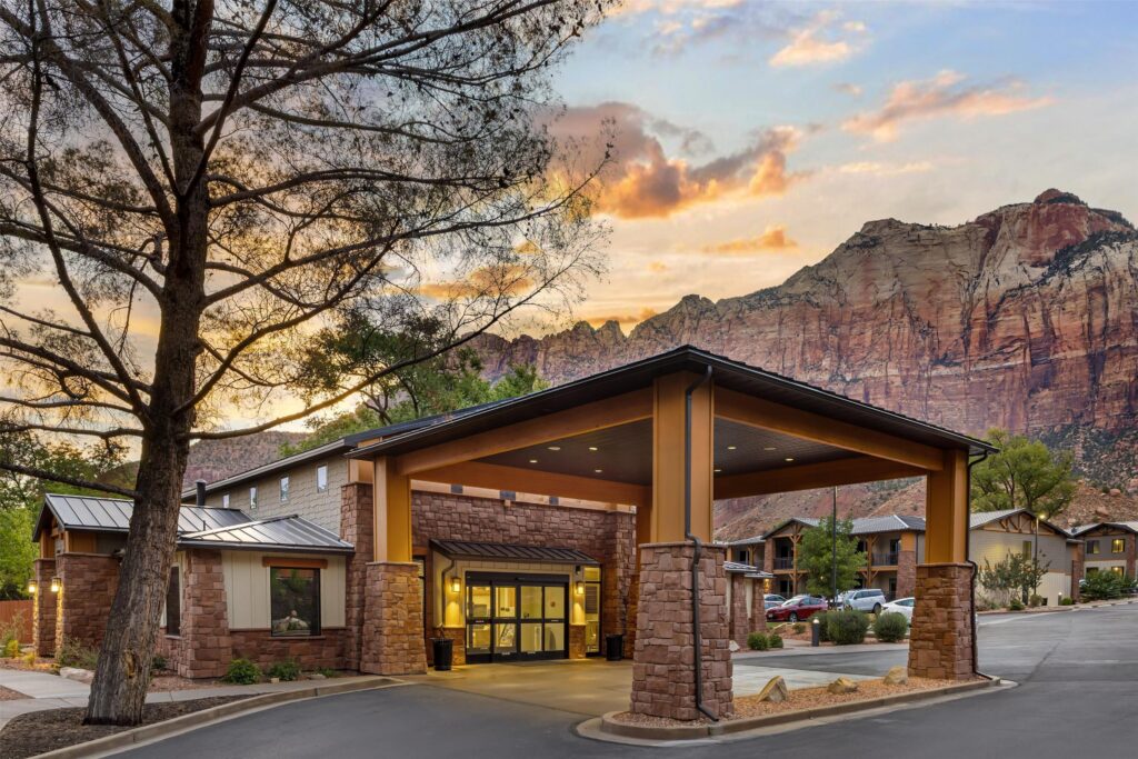 Best Western Plus in Springdale, Utah, at dusk, with its illuminated stone-and-timber porte-cochère entrance glowing warmly against the dramatic red sandstone cliffs of Zion beneath a sunset sky.