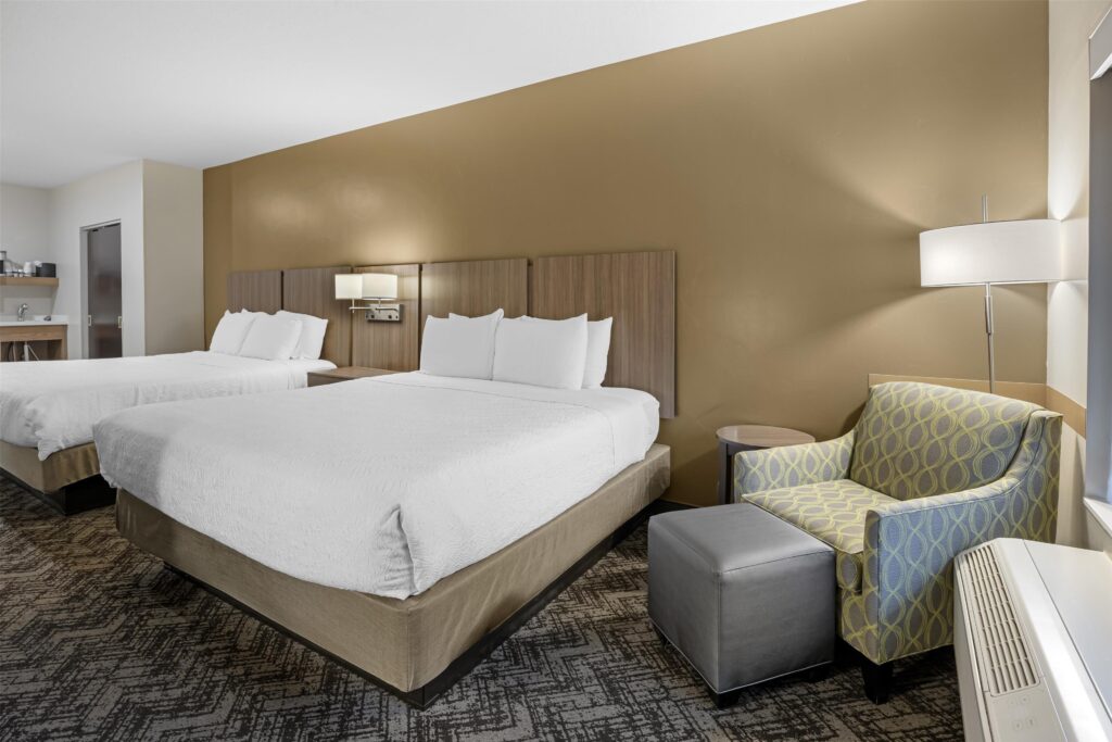 Best Western Plus guest room in Springdale, UT with two queen beds, white bedding, wood-panel headboards against a gold accent wall, and a blue-and-green armchair with ottoman.