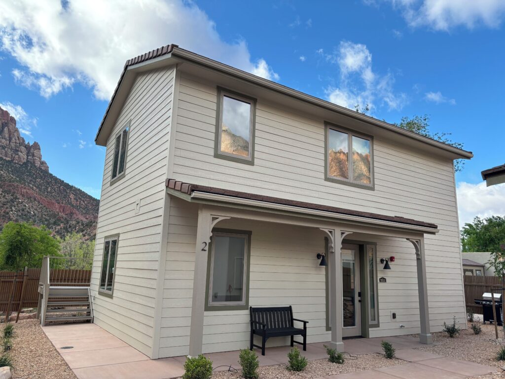 Cottages at Worthington Gallery in Springdale, a two-story cream lap-siding cottage with a covered porch, framed by Zion's red sandstone cliffs rising behind it under a partly cloudy sky.
