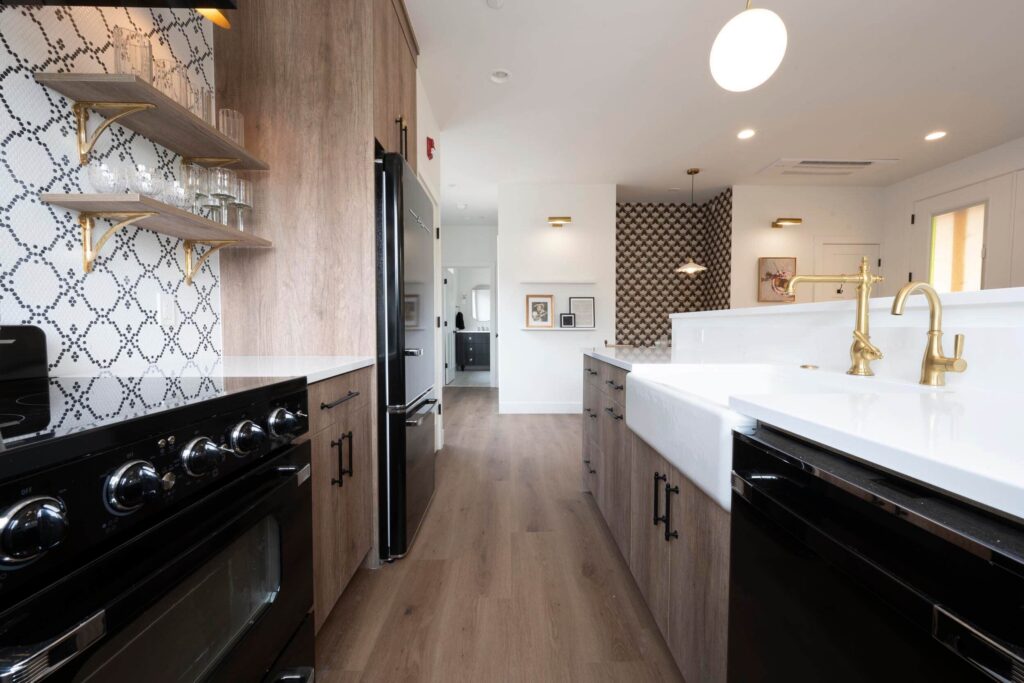 Galley kitchen inside the Cottages at Worthington Gallery in Springdale, with black appliances, wood-tone cabinetry, a white farmhouse sink with brass faucets, patterned tile backsplash, and warm hardwood floors.