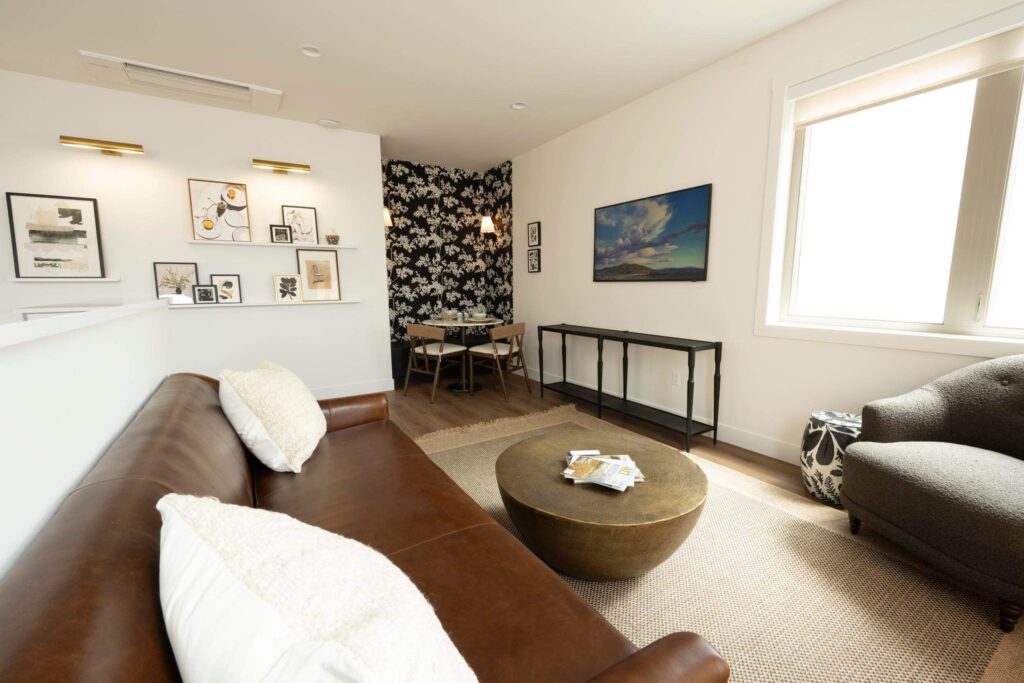 Bright open-plan living room with a brown leather sofa, brass drum coffee table, and gray tufted chair, flowing into a dining nook with bold black-and-white floral wallpaper and natural light throughout.