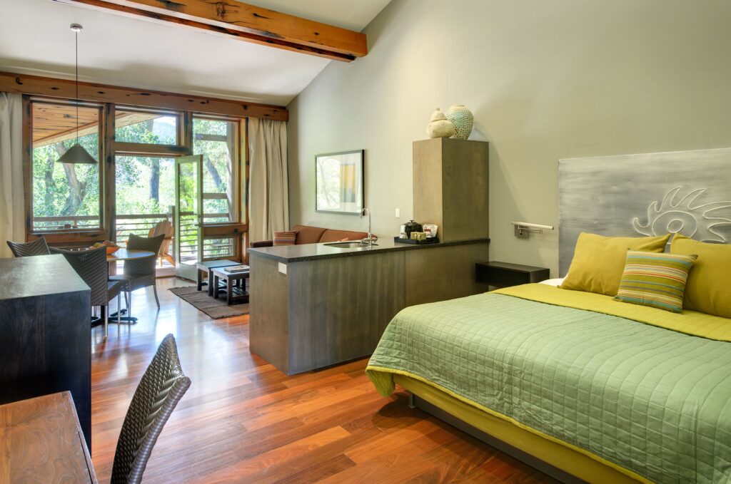 Desert Pearl Inn studio room in Springdale, Utah, with a king bed, ornate silver headboard, green quilted bedding, kitchenette, and glass doors opening to a tree-lined balcony.