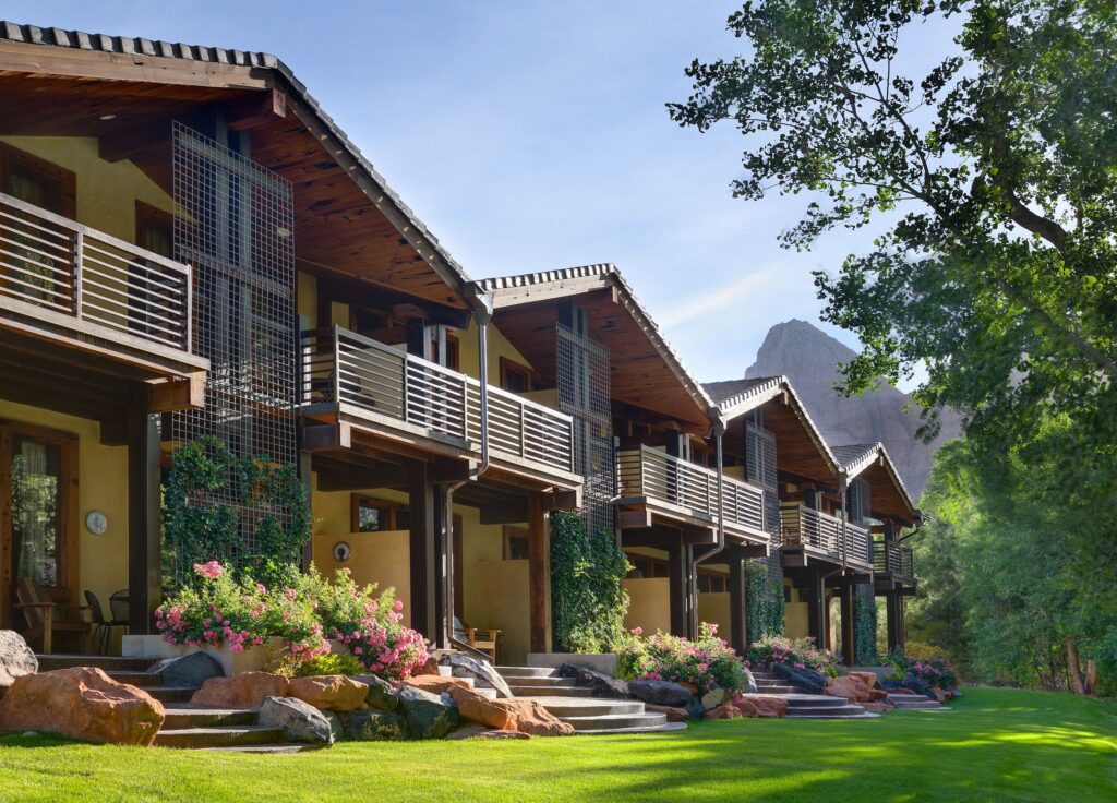 Desert Pearl Inn guest buildings in Springdale, Utah, with yellow stucco walls, wood-beamed gabled roofs, and ivy-draped balconies overlooking a lawn with roses and sandstone boulders, red rock peaks visible behind.