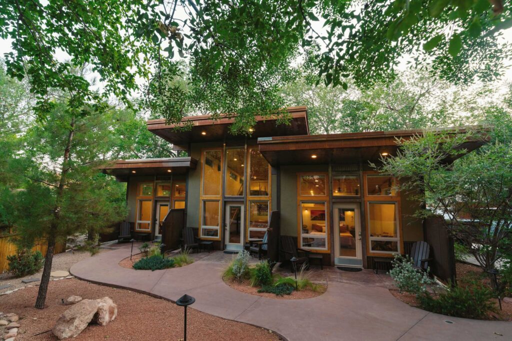 Nama-Stay lodging in Springdale, Utah at dusk, its modern low-profile structure glowing warmly through floor-to-ceiling windows beneath a canopy of mature trees, with a curved concrete path leading to the entrance.