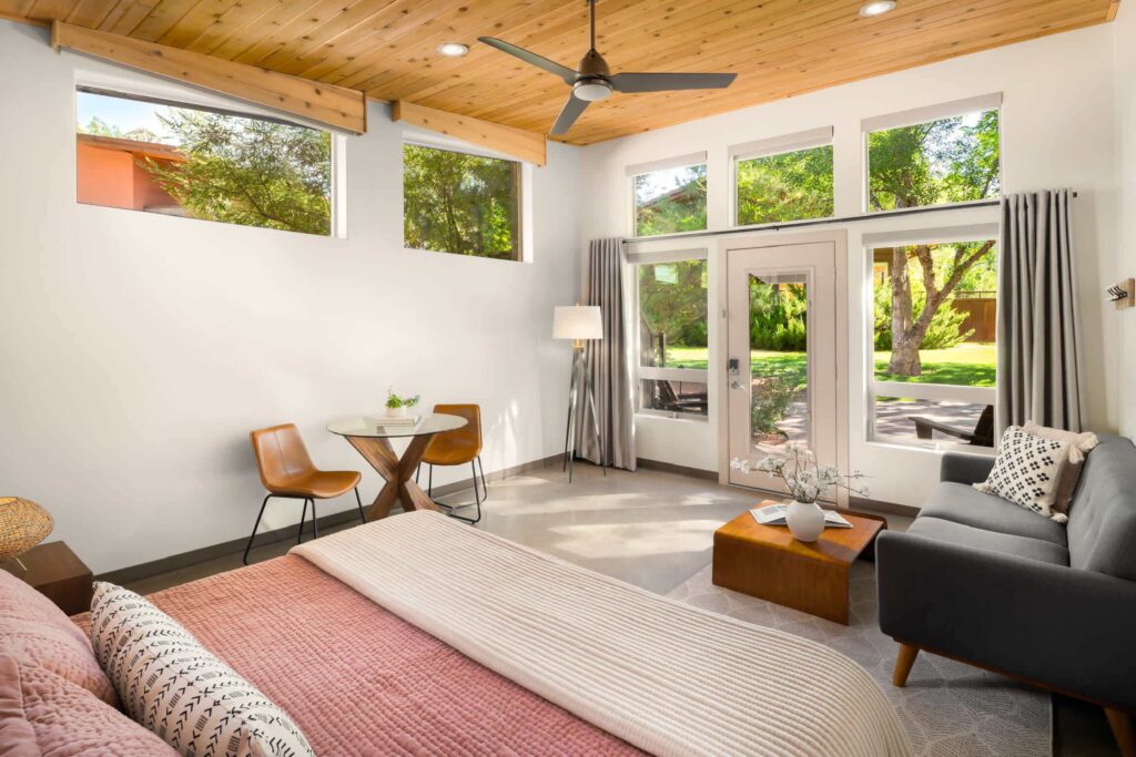 Bright studio room at Nama-Stay in Springdale, with a king bed in pink bedding, leather chairs at a bistro table, wood plank ceiling, and windowed door opening to a lush green garden.