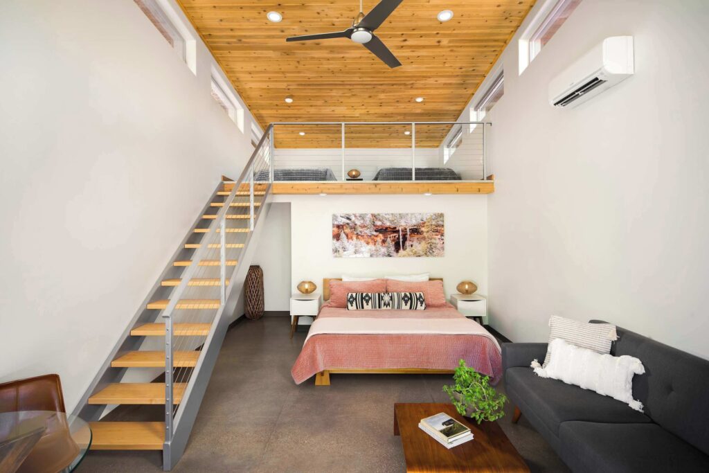 Interior of a Nama-Stay studio in Springdale, UT, with a king bed, floating steel-and-wood staircase to a mezzanine loft, wood-planked ceiling with fan, and modern furnishings throughout.