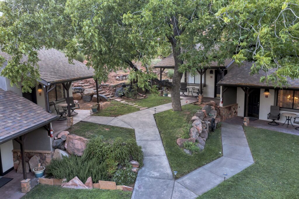 Aerial view of Red Rock Inn's courtyard in Springdale, UT, with forking walkways, manicured grass, shade trees, red sandstone boulders, and cottage units with covered patios arranged around a central garden.