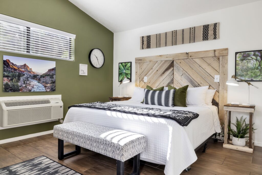 Guest room at Red Rock Inn in Springdale, UT with a king bed, reclaimed wood chevron headboard, white quilted bedding, and an olive-green accent wall featuring a red rock canyon photograph.