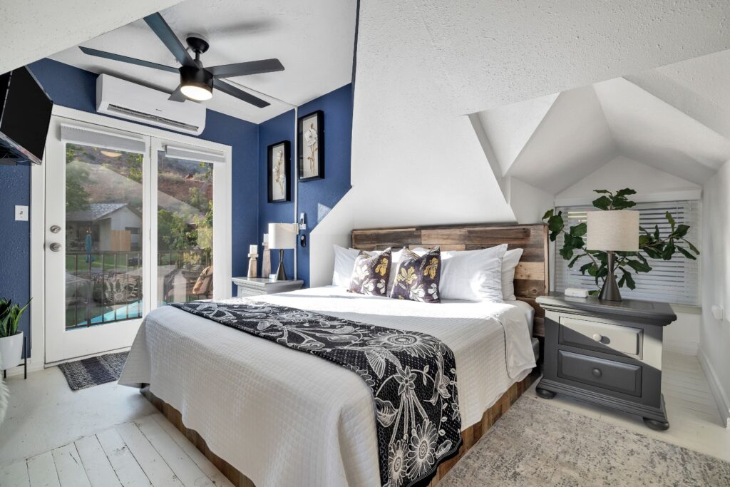 King bedroom at Red Rock Inn in Springdale, Utah, with a navy accent wall, reclaimed wood headboard, white quilt, and French doors opening to a view of surrounding hills.