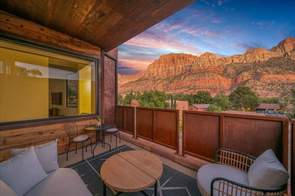 Furnished private balcony at Zion Sage Suites with lounge chairs and bistro table, backed by a dramatic sunset over Springdale's towering red sandstone cliffs.