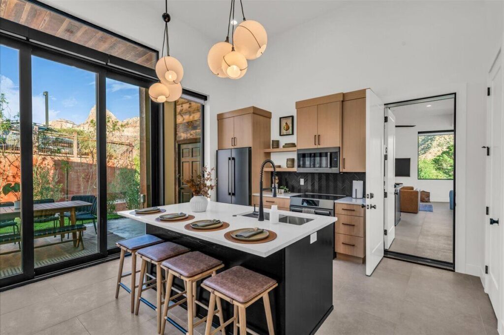 Zion Sage Suites kitchen in Springdale, UT, with a quartz island set for four, warm wood cabinetry, herringbone tile backsplash, and sliding glass doors opening to a patio with red rock views beyond.