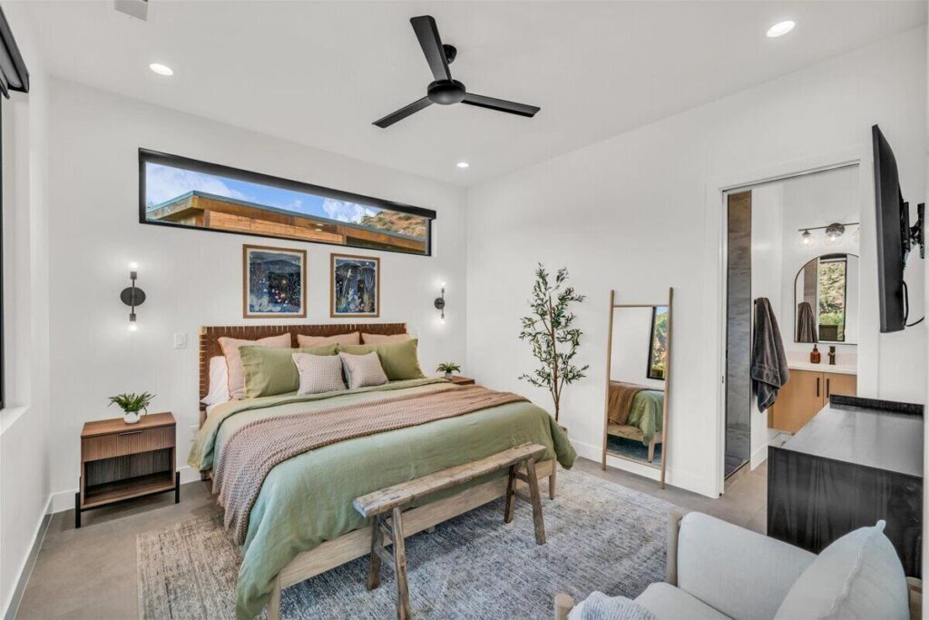 Primary bedroom at Zion Sage Suites in Springdale, with a queen bed in sage green linen, woven leather headboard, clerestory window, and an open doorway revealing the en suite bathroom.
