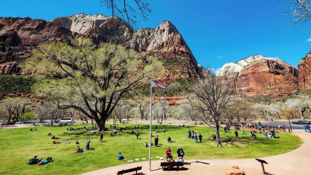 Crowds relaxing on the broad lawn in front of Zion Lodge, with a flagpole at center, shading trees, and dramatic red sandstone canyon walls rising under a vivid blue sky.