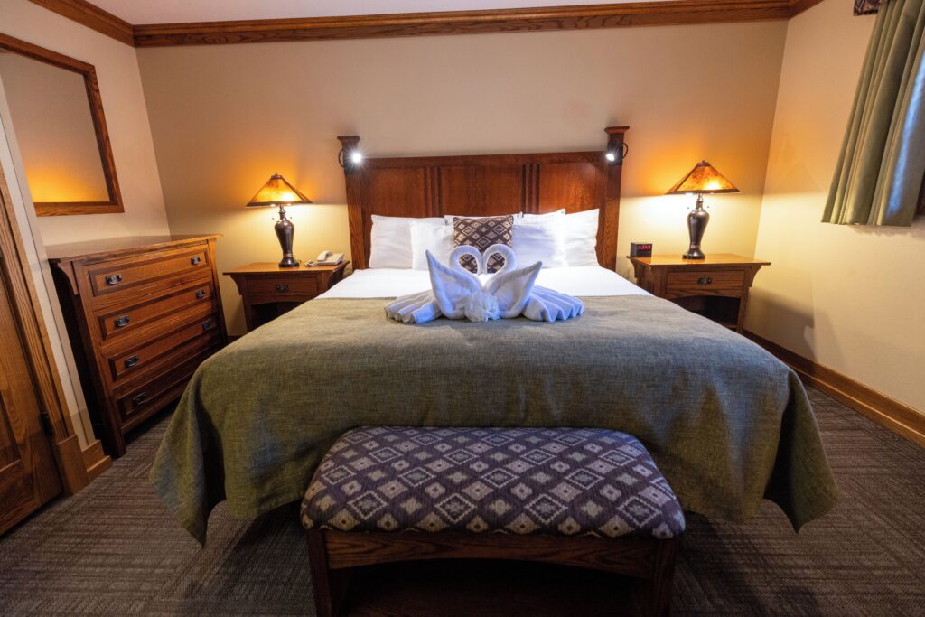 Zion Lodge guest room with a king bed dressed in a green coverlet and two towels folded into swans, flanked by craftsman-style nightstands with amber lamps and a dark wood headboard with reading lights.