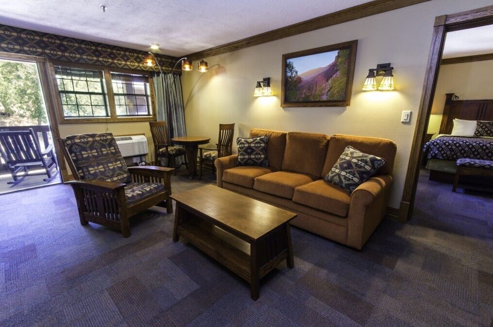 Zion Lodge suite interior with Southwest-patterned upholstery, canyon landscape painting, and a sliding glass door opening to a porch with rocking chairs among the trees.