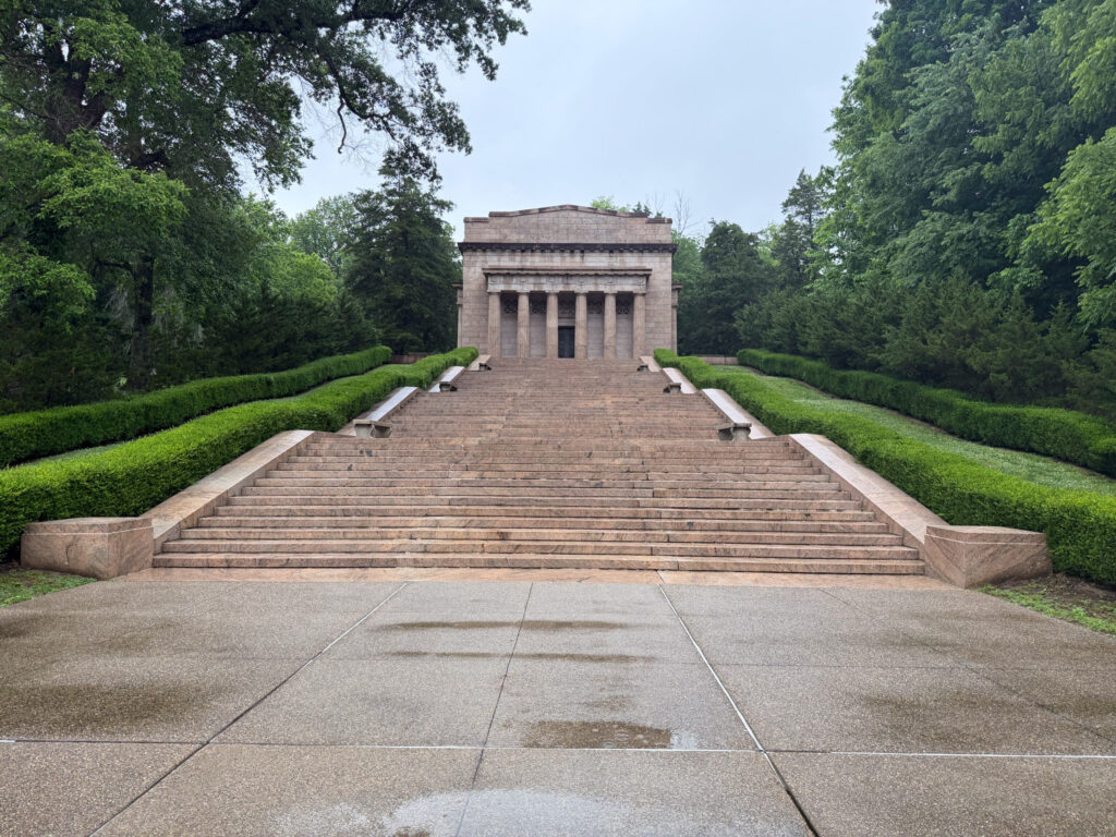 The neoclassical granite memorial building at Abraham Lincoln Birthplace's Birthplace Unit, its columned facade rising above a long symmetrical staircase flanked by trimmed hedgerows and tall trees under an overcast sky.
