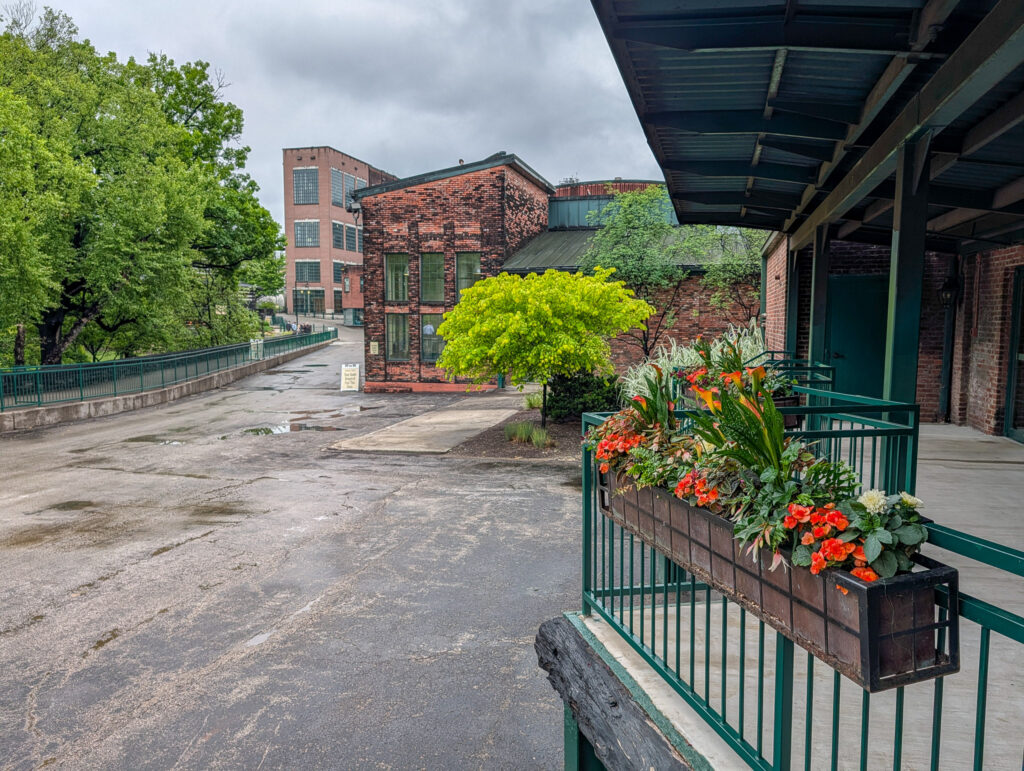 View from a covered porch at Buffalo Trace Distillery, looking out over a wet parking lot toward red brick distillery buildings and lush trees under an overcast sky, with orange and red flowers in a planter along the railing.