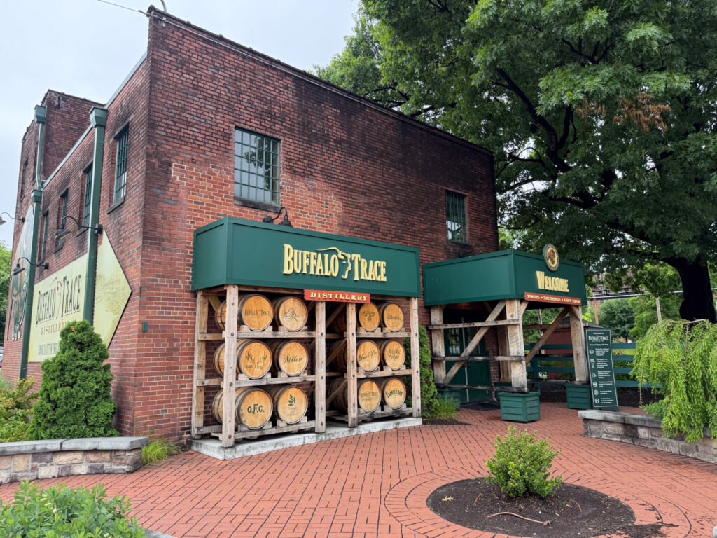 Visitor entrance to Buffalo Trace Distillery in Kentucky, with a green-canopied welcome archway and a wooden barrel display featuring O.F.C., Weller, and Buffalo Trace branded barrels against the red brick facade.