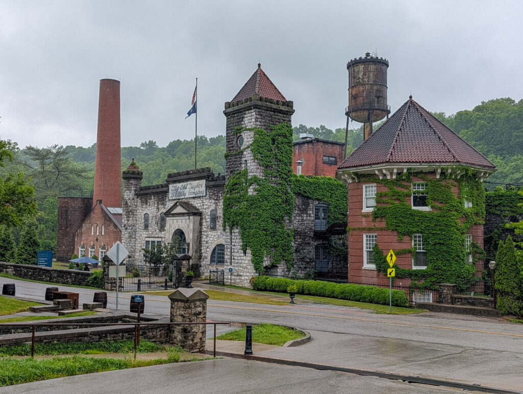 Castle + Key distillery entrance on an overcast day, showing the ivy-draped stone gatehouse with conical turret, a brick smokestack, wooden water tower, and a sign reading 'The Old Taylor Distillery Company' on the facade.