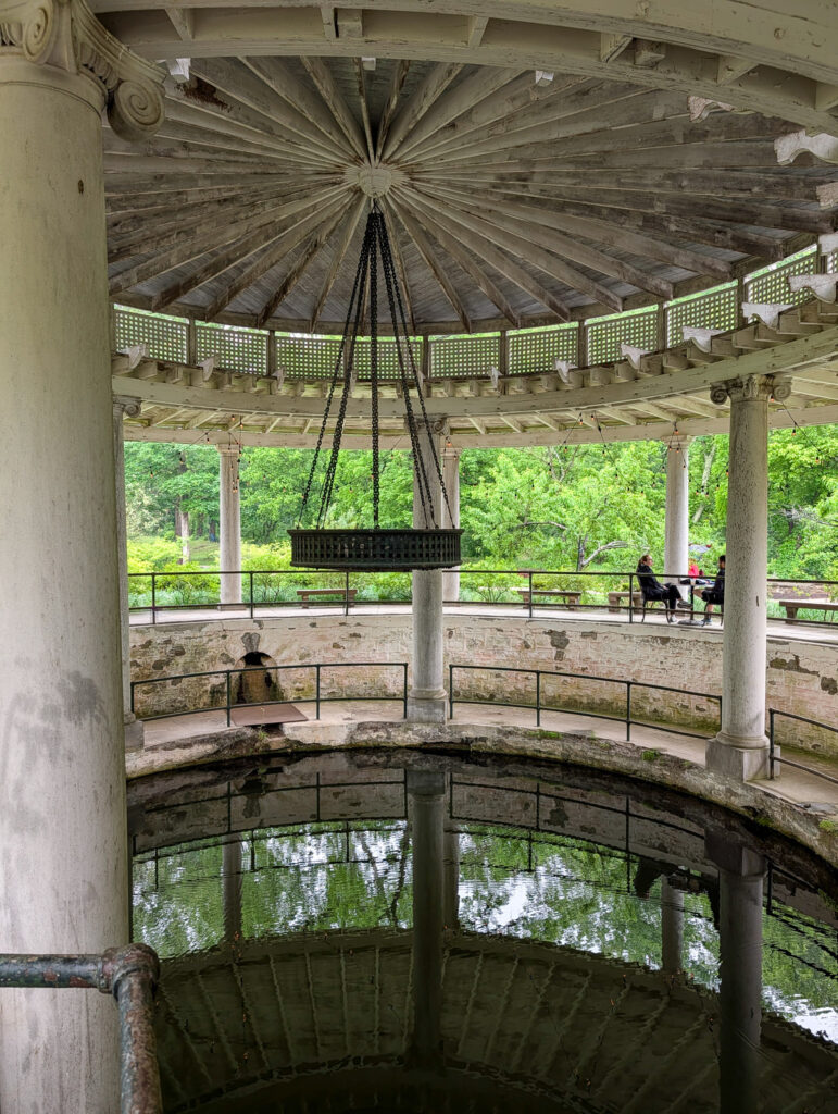 The historic spring house at Castle and Key distillery in Kentucky, with white Ionic columns supporting a radial wooden ceiling and chandelier, surrounding a sunken circular pool of still green water open to lush trees beyond.
