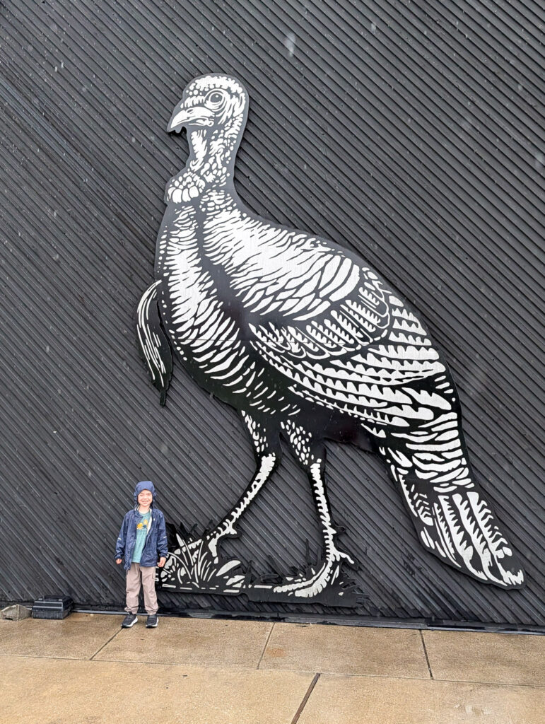 A young child stands beside a massive black-and-white metal wild turkey cutout at Wild Turkey distillery in Kentucky, the towering artwork dramatically dwarfing the child in scale.