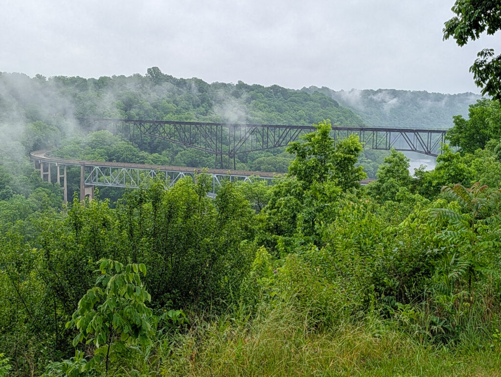 Two steel truss bridges span a wooded river gorge near Wild Turkey on the Kentucky Bourbon Trail—a curved road bridge and a taller railroad trestle crossing in parallel beneath misty, overcast skies.