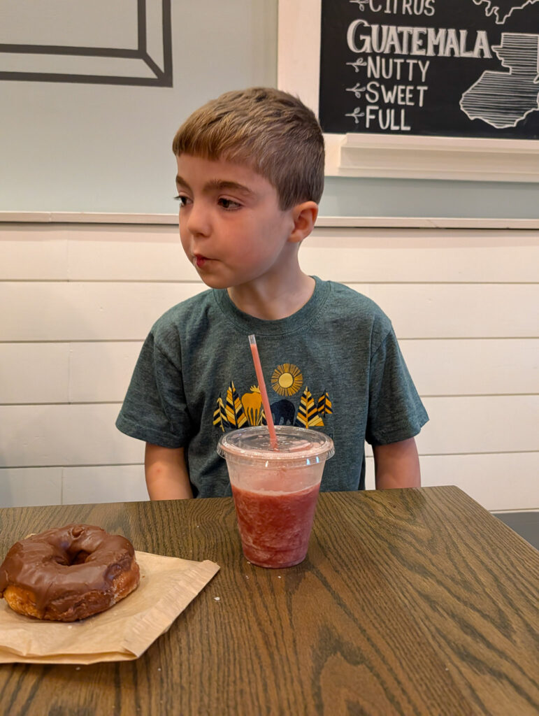 A young boy sits at a wooden table inside Old School Coffee in Lexington, KY, with a chocolate-glazed donut and a red blended drink, a chalkboard menu visible behind him.